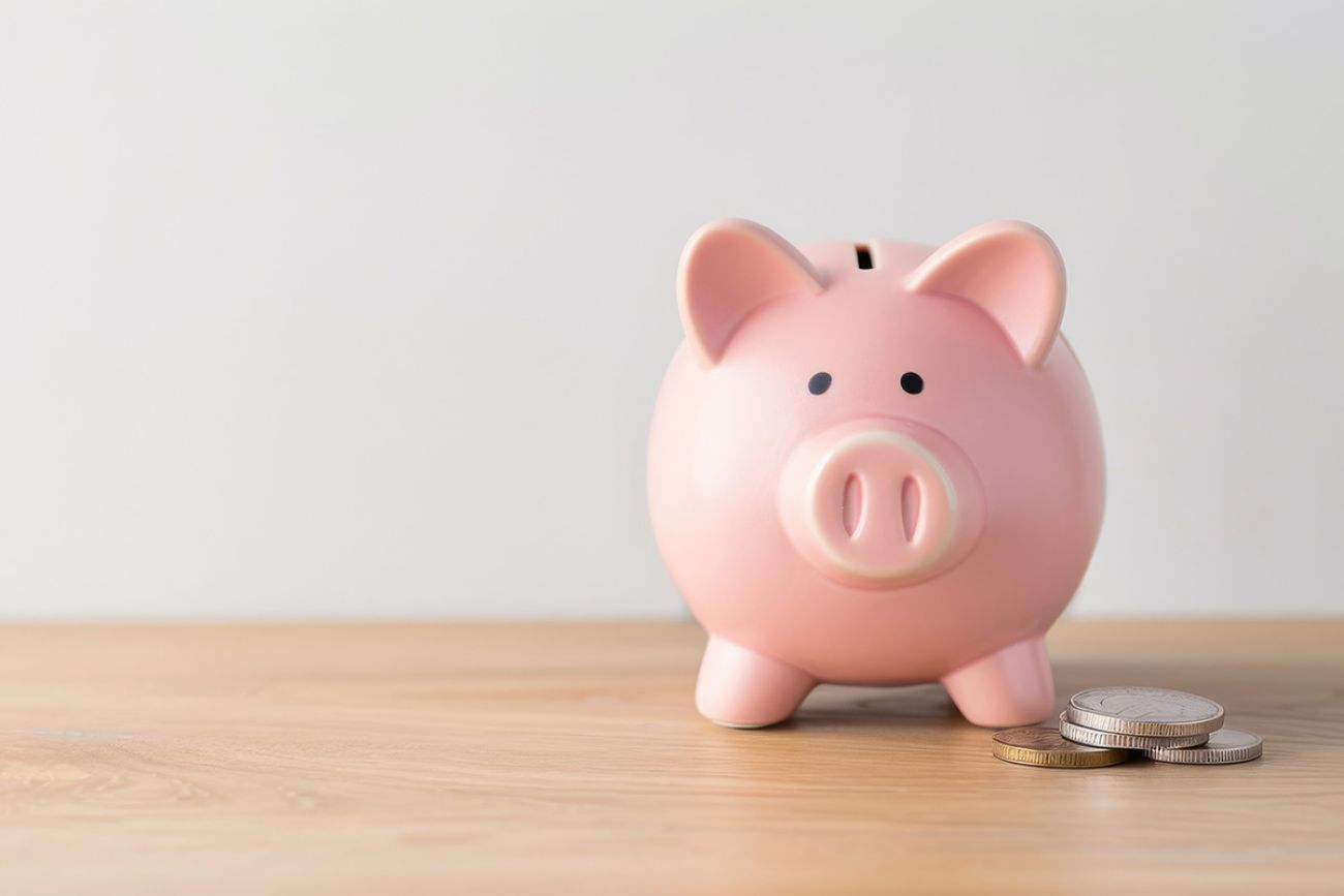 A pink piggy bank overflowing with coins is placed on a wooden table, against a white wall background.