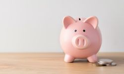 A pink piggy bank overflowing with coins is placed on a wooden table, against a white wall background.